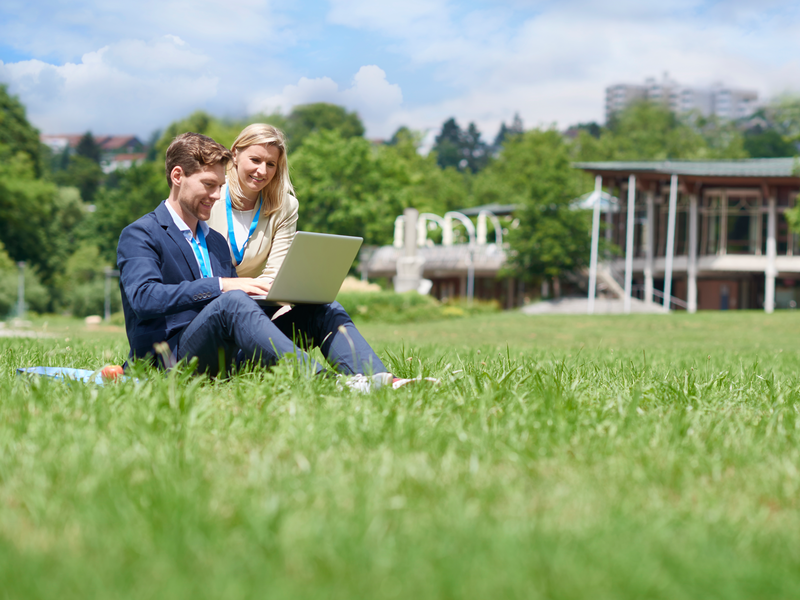 Ein Mann und eine Frau sitzen auf einer Wiese und benutzen zusammen einen Laptop, mit einem Gebäude und Bäumen im Hintergrund.