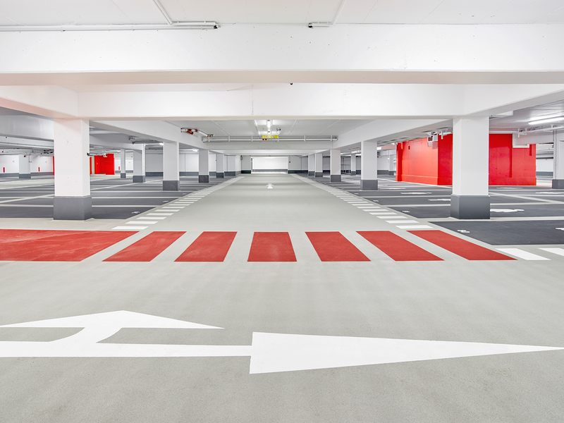 Ein leerer, gut beleuchteter Parkhausbereich mit weißen Pfeilen, weißen Markierungen und roten Abschnitten an den Wänden und am Boden.