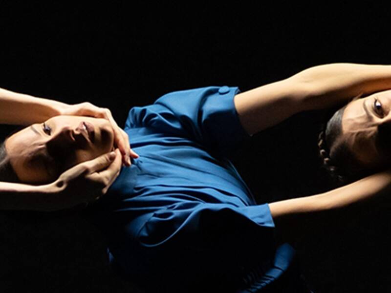 Drei Tänzerinnen in blauen Kleidern sind in einer künstlerischen Pose miteinander verbunden; sie knien auf dem Boden und strecken ihre Arme elegant aus.