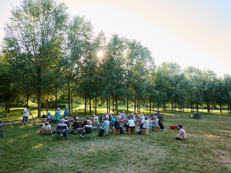 Eine Gruppe von Menschen sitzt bei sonnigem Wetter in einem Park auf Klappstühlen und hört einem Musiker zu, der ein Didgeridoo spielt, während im Hintergrund Bäume zu sehen sind.