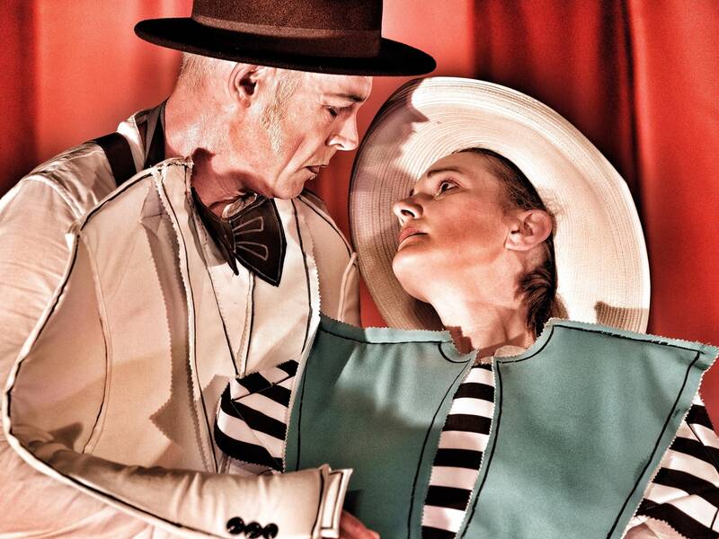 Ein Mann und eine Frau in vintage-inspirierter Kleidung und Hüten schauen sich intensiv vor einem roten Vorhang an.