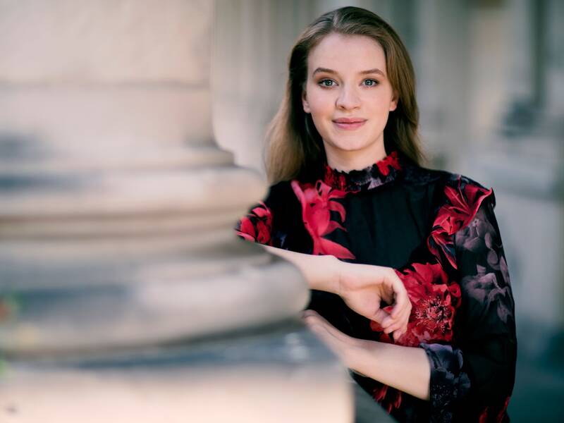 Eine lächelnde Person mit langen Haaren trägt ein schwarzes Kleid mit roten Blumenmustern und lehnt mit den Armen auf einer verschwommenen Marmorsäule.