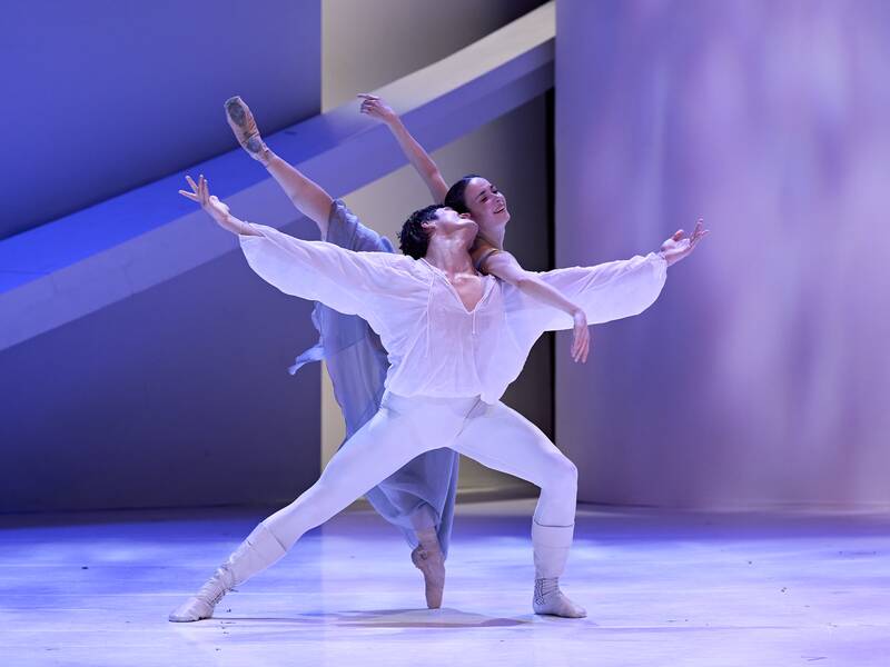 Zwei Balletttänzer in fließenden weißen Kostümen führen eine elegante Hebefigur auf einer Bühne mit blau-violetter Beleuchtung aus.