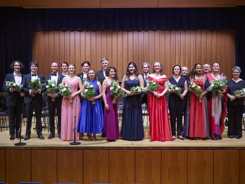Eine Gruppe von elegant gekleideten Personen steht auf einer Bühne und hält Blumensträuße in den Händen.