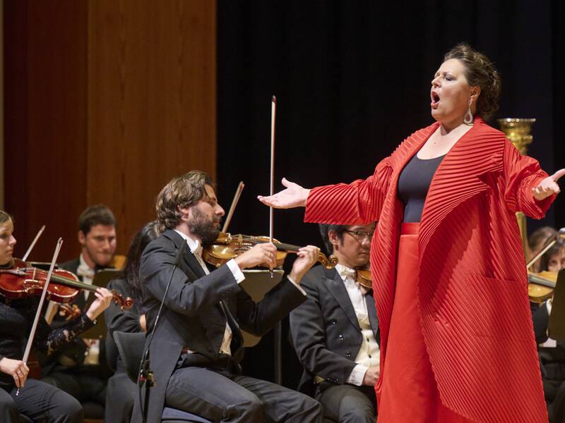 Eine Frau mit rotem Mantel und rotem Rock singt kraftvoll, im Hintergrund spielt ein Orchester