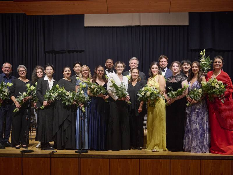Ein Gruppenbild auf einer Bühne mit 19 Personen, die fröhlich und lächelnd Blumensträuße in den Armen halten