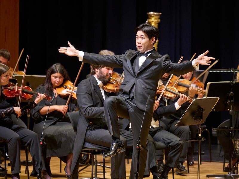 Im Vordergrund singt ein Mann und hebt dabei seinen rechten Bein hoch, während im Hintergrund ein Orchester spielt