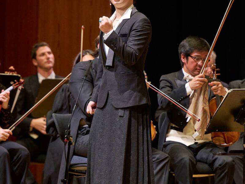 Ein Mann lächelt im Vordergrund und hält ein Faust geballt vor sich, während im Hintergrund ein Orchester spielt