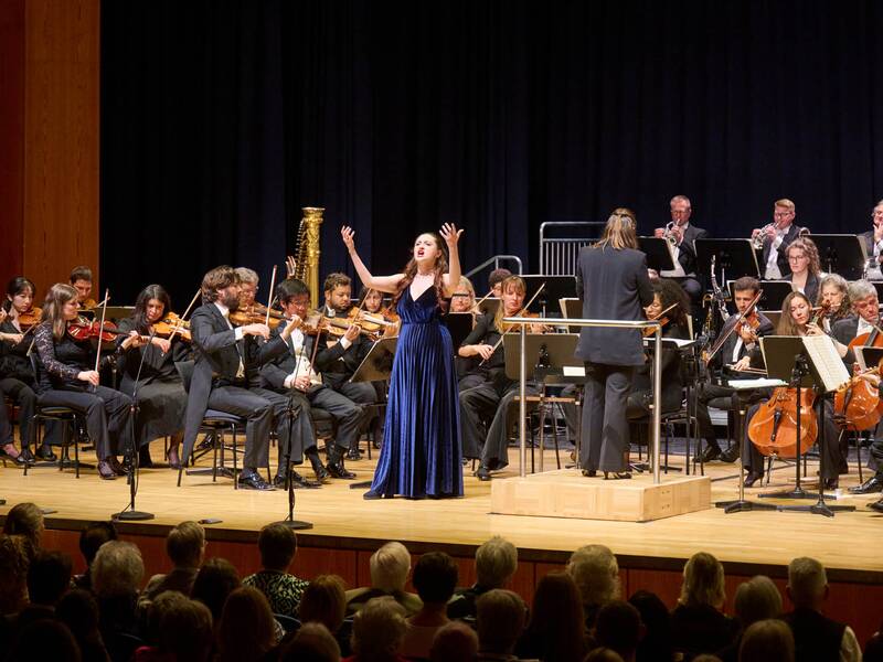 Eine Frau im blauen Kleid singt und hebt die Hände in die Richtung Himmel, während ein Orchester hinter ihr spielt