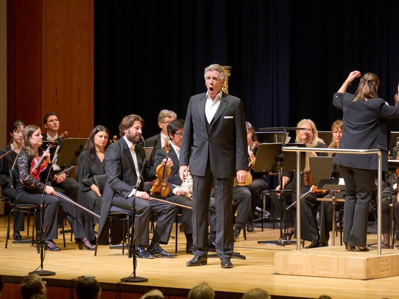 Ein Mann singt kraftvoll eine Arie, hinter ihm spielt das Orchester, das ihn begleitet