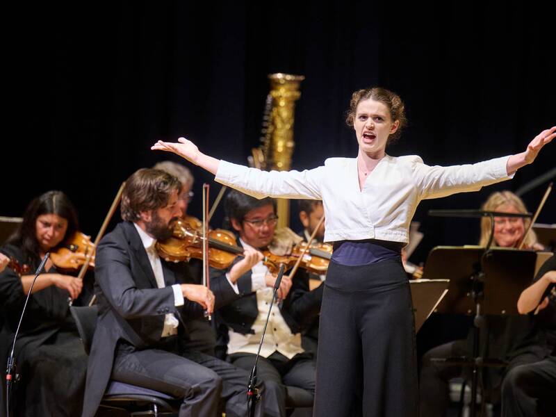 Eine junge Dame mit weißer Bluse und schwarzer Hose singt eine Arie und streckt ihre Arme aus