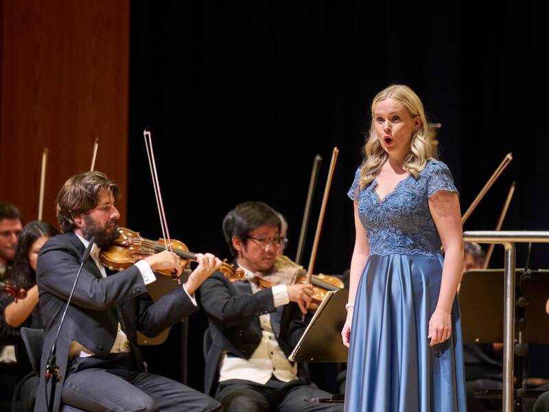Eine junge Dame im blauen Abendkleid singt eine Arie, im Hintergrund kann man das Orchester spielen sehen