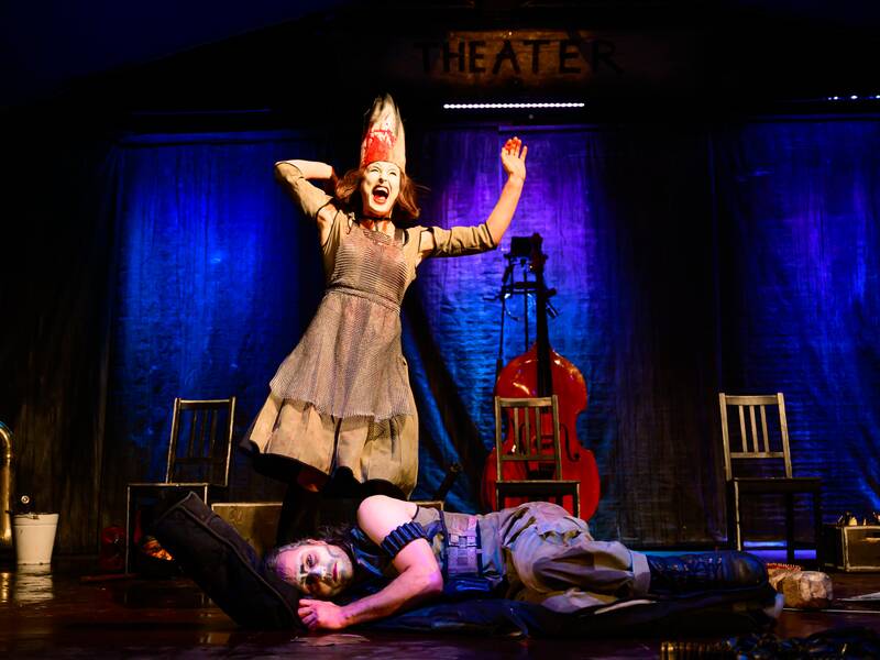 Eine Frau auf einer Theaterbühne hat die Arme erhoben und einen spitzen Hut auf, während eine andere Person am Boden liegt und ein Cello im Hintergrund steht.