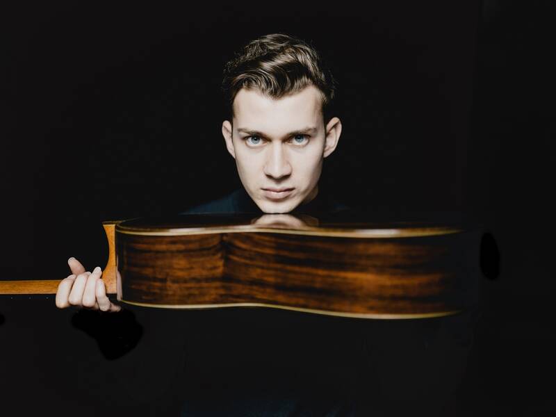 Ein Mann mit ernstem Gesichtsausdruck hält eine Gitarre quer vor sich in einem dunklen Hintergrund.