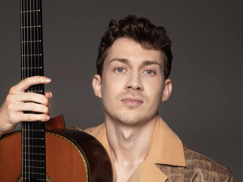 Ein Mann in einem braunen Lederjackett hält eine Gitarre vor einem neutralen Hintergrund.